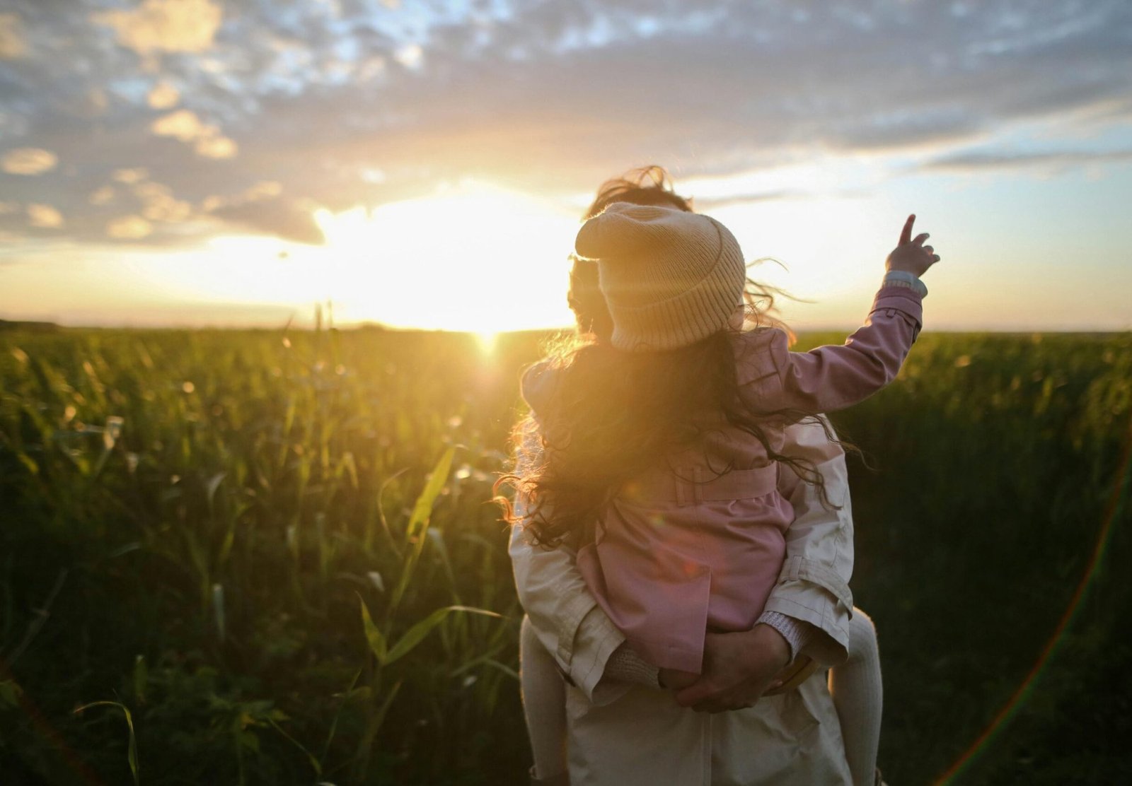 A mother and daughter embrace and point at the sunset in a grassy field.